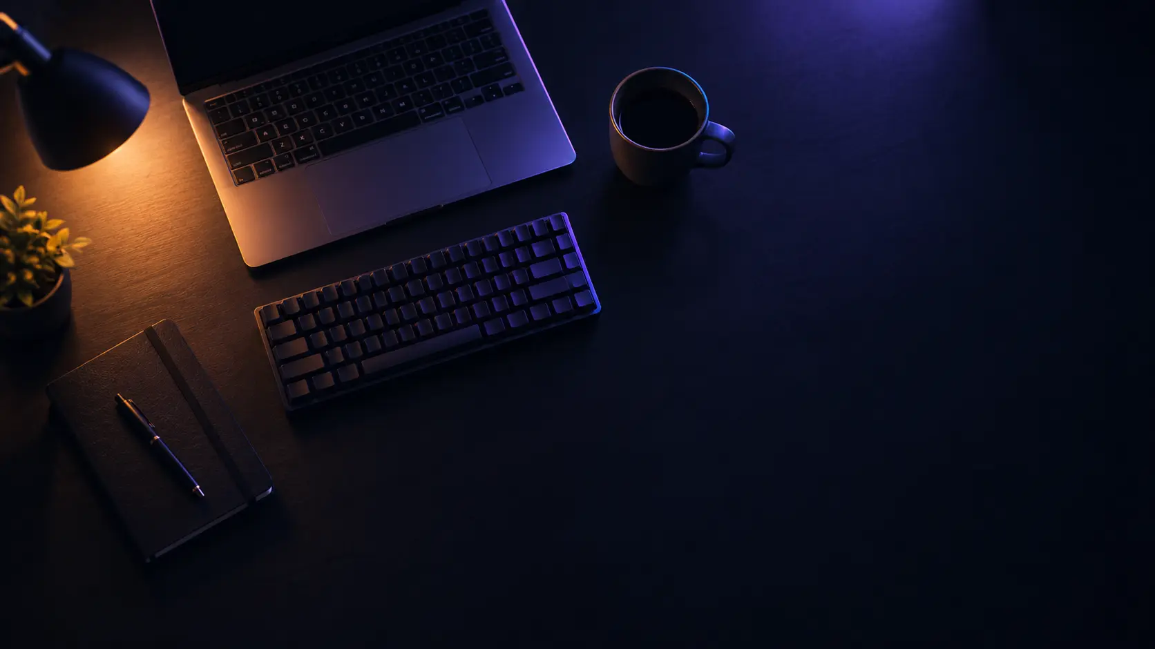 Biurko widziane z góry: laptop, klawiatura, notatnik i kubek w świetle lampy